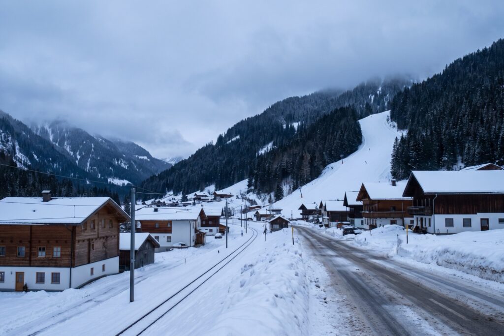 A single railway track and a cleared road running side by side through a snow-covered valley village of timber chalets, with a ski slope coming down through the forest behind