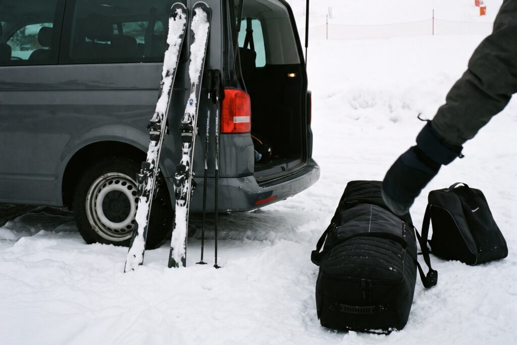 Two pairs of skis and poles leaning against a gray transfer van with its tailgate open, a duffel bag and a boot bag on the snow beside it