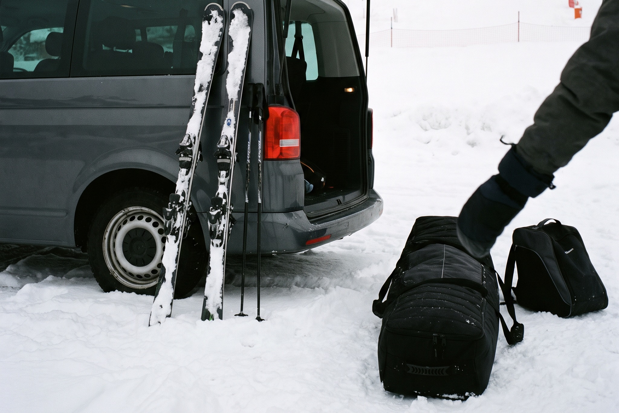 Two pairs of skis and poles leaning against a gray transfer van with its tailgate open, a duffel bag and a boot bag on the snow beside it