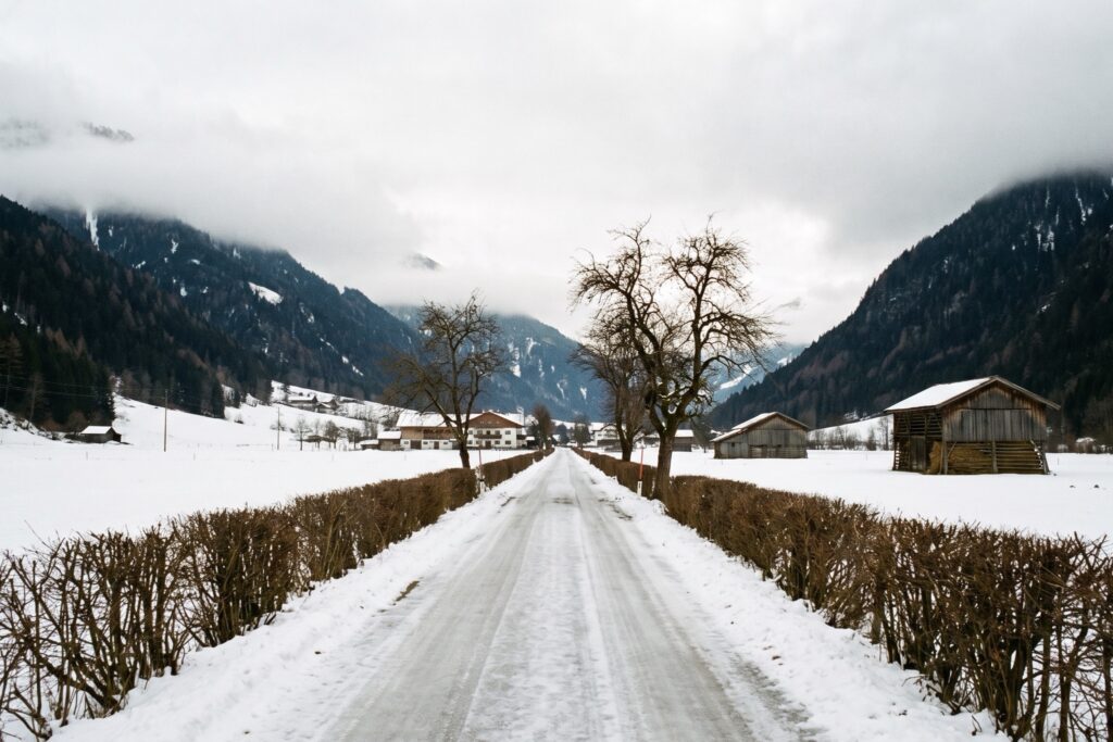 A snow-covered lane between bare hedges running up a narrow winter valley, past timber barns and farmhouses, with forested slopes and low cloud on both sides