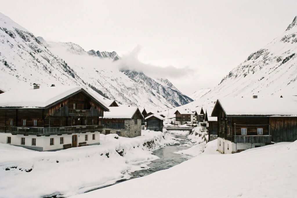 Old timber houses under deep snow on both banks of a mountain stream, in a narrow high valley with snowy slopes rising on either side and cloud on the peaks
